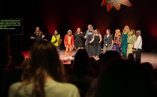 A group of 11 people stand on stage smiling. There is are the backs of audience members standing up in the foreground, and a sign on stage saying WOW Women of the World
