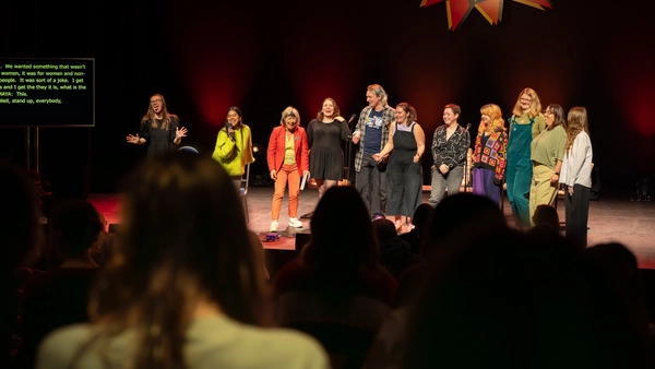 A group of 11 people stand on stage smiling. There is are the backs of audience members standing up in the foreground, and a sign on stage saying WOW Women of the World