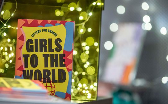 A colourful book titled "Girls to the World" sits amidst soft glowing lights.