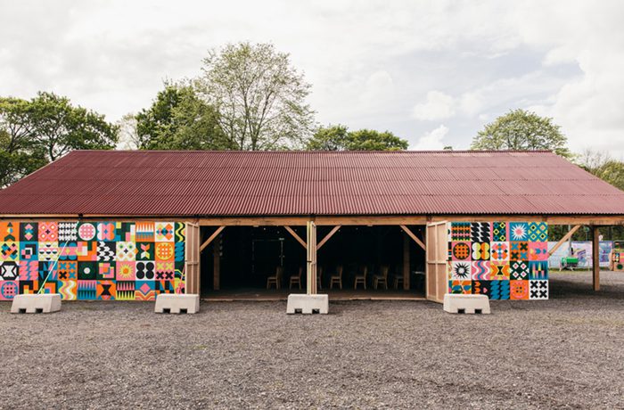 A vibrant wooden barn with a red roof and colourful geometric patterns on the walls, surround by trees and a cloudy sky