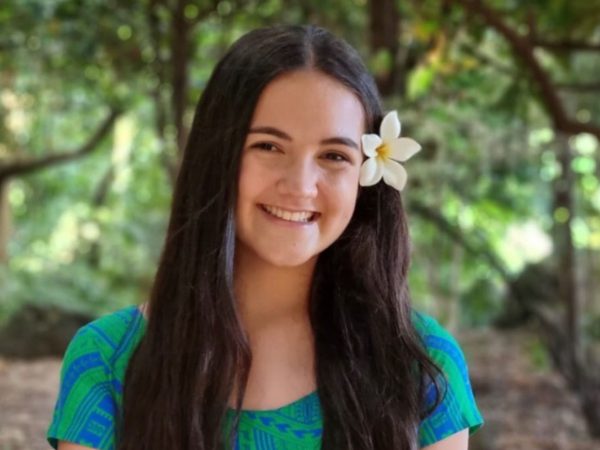 A young woman with long dark hair smiles warmly. She has a white flower in her hair. The backdrop is a sunlit forest.