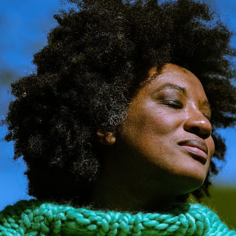 Aja Barber has her eyes closed and her face bathed in light. She is wearing a green knitted jumper, and is against a blue background.