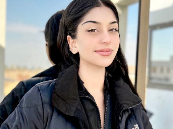 A young woman with long dark hair smiles slightly. She stands outside against a window on a sunny day.