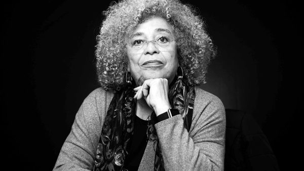 A black and white headshot of Angela Davis, a woman with grey afro hair and glasses, she is sat down with her hand on her chin.