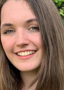 Young woman with long brown hair smiles warmly, standing in front of green ivy leaves