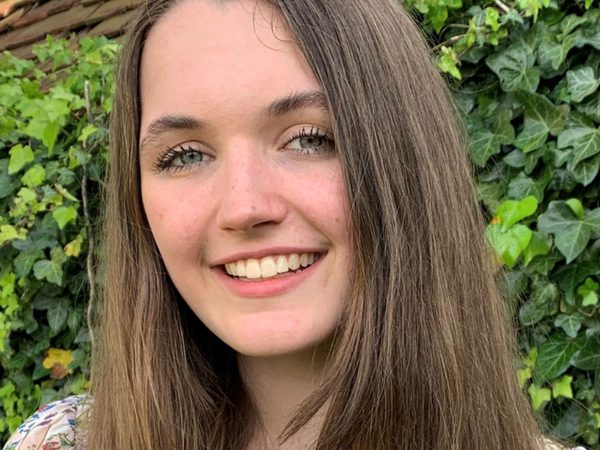 Young woman with long brown hair smiles warmly, standing in front of green ivy leaves