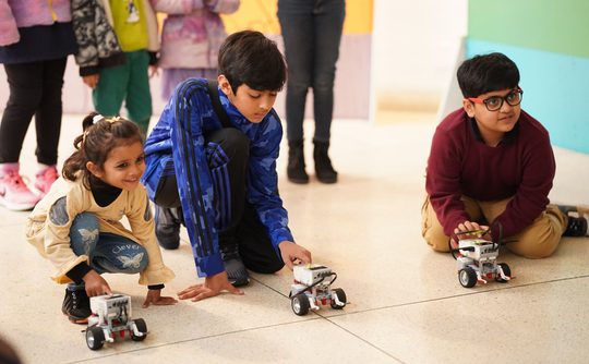 Three children, two boys and a girl, crouch on the floor with their fingers pressed on small robots with wheels.