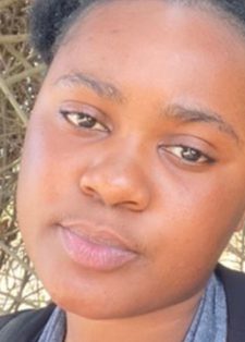A young woman with a neutral expression stands outdoors, framed by tangled branches in the background