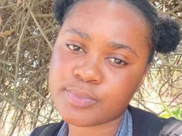 A young woman with a neutral expression stands outdoors, framed by tangled branches in the background