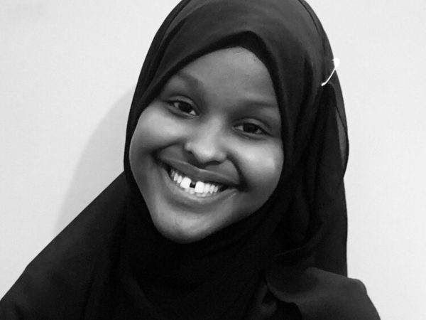 Black and white image of a young woman smiling wearing a dark hijab against a light background.