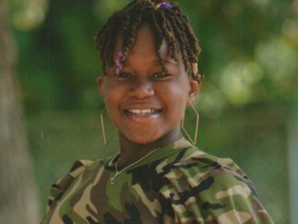 Smiling individual with short braided hair, wearing a camo-patterned shirt and hoop earrings, stands outdoors with blurred green foliage in the background.