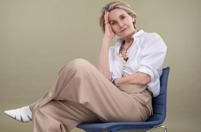 Mary Portas sits on a blue chair with her legs crossed. Her head is rested in her hand, and she is wearing a smart white shirt, loose brown trousers and pointed white shoes.
