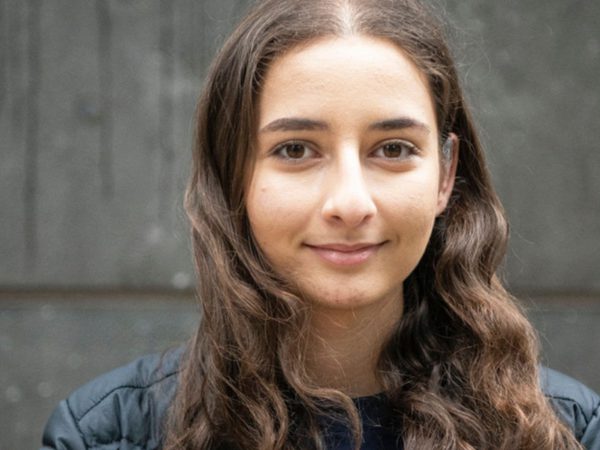 Young woman with long wavy hair, wearing a black jacket, stands against a grey concrete wall smiling subtly