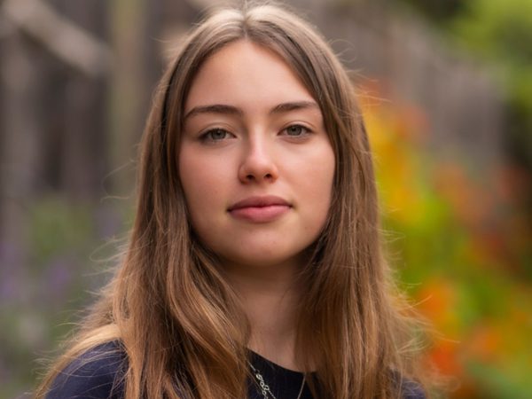 A young woman with long brown hair stands outdoors, looking confidently at the camera