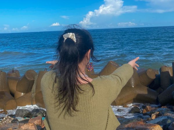 A woman with long dark hair in a ponytail, wearing a green sweater, faces the ocean. She gestures to the sea, surrounded by large rocks