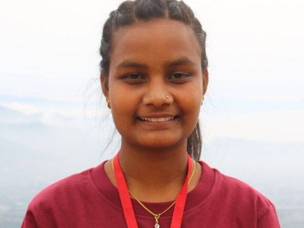 Saru Khadka looks into the camera smiling. She has long brown hair tied up in a pony tail, and is wearing a red top and lanyard.