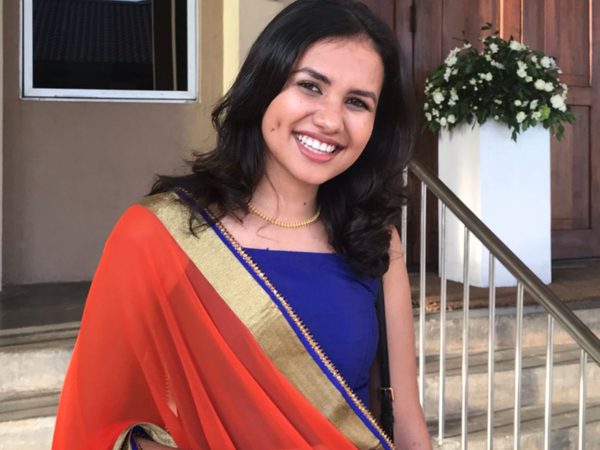 A woman smiles warmly, wearing a vibrant orange and gold sari with a blue blouse