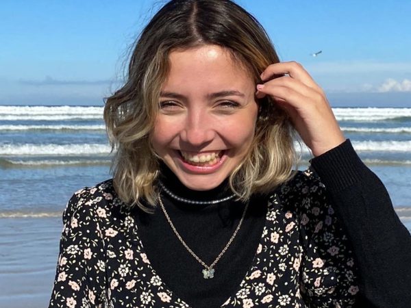 Young woman with a beaming smile stands on a beach. She wears a floral dress over a black turtleneck