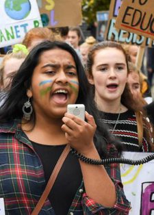 A diverse group of young protesters march, holding climate change signs. A girl with face paint speaks into a megaphone,