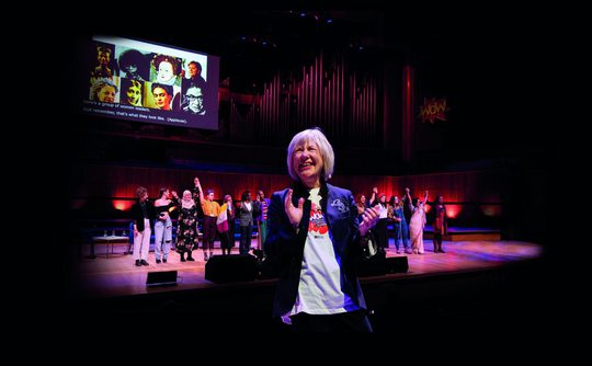 Jude Kelly smiles in front of a stage, clapping her hands together. There is a screen and a group of people behind her on the stage, which is lit with pink and blue lighting.