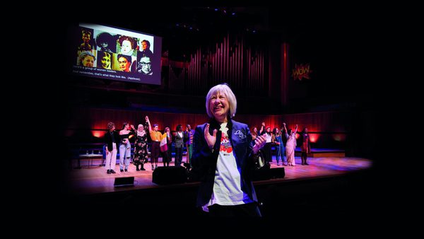 Jude Kelly smiles in front of a stage, clapping her hands together. There is a screen and a group of people behind her on the stage, which is lit with pink and blue lighting.