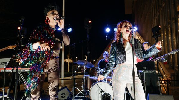 Two singers perform on an outdoor stage with a band behind them. They are wearing elaborate shiny outfits.