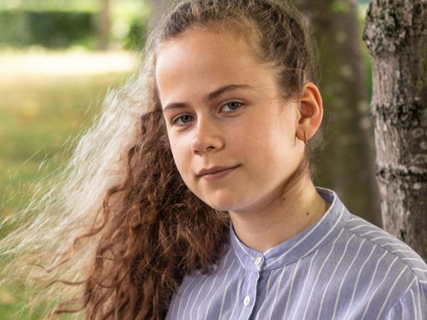 A young person with long curly hair stands by a tree, wearing a blue striped shirt