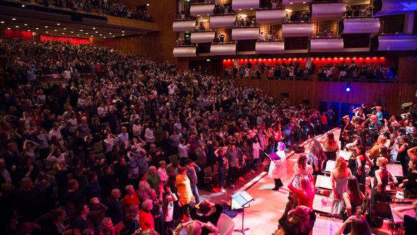A vibrant concert hall is filled with a standing audience, applauding enthusiastically. The stage is lit warmly, showcasing musicians and performers.