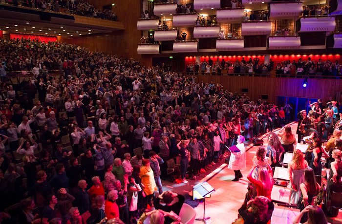 A vibrant concert hall is filled with a standing audience, applauding enthusiastically. The stage is lit warmly, showcasing musicians and performers.