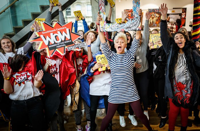 A diverse group of people celebrating energetically indoors. They hold colourful signs expressing joy and excitement.