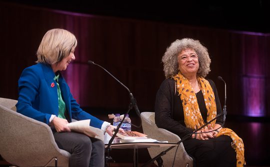 Two women are seated on stage in conversation. One wears a blue blazer, the other a yellow scarf. Both appear engaged, with a warm atmosphere.