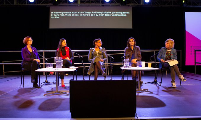Five women sit on a stage with microphones in front of them looking pensive and deep in conversation