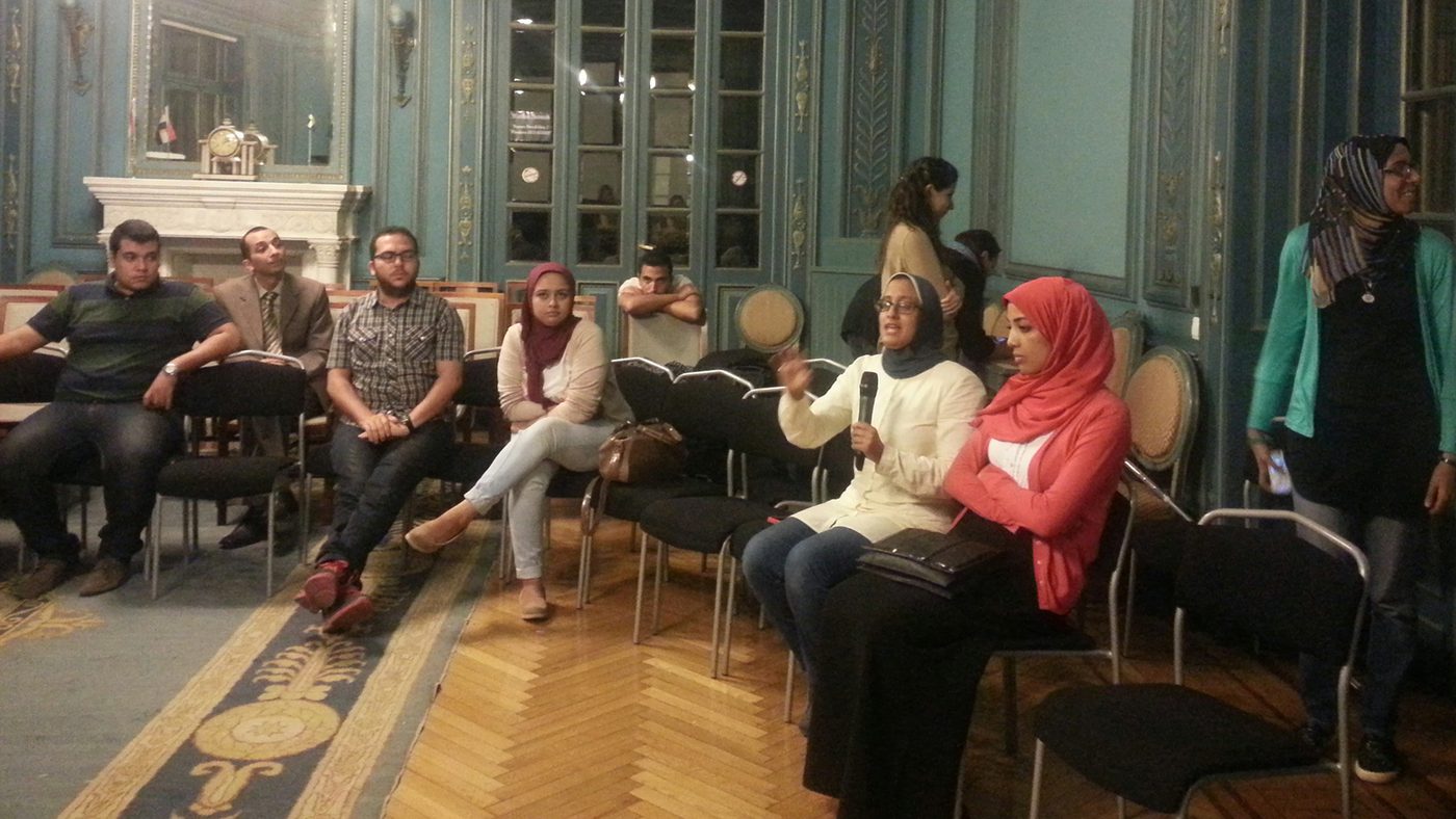 A group of people sat down in a large historical room with big windows, gold details on the walls. They are sat in a semi-circle, and one person is speaking and is holding a microphone.