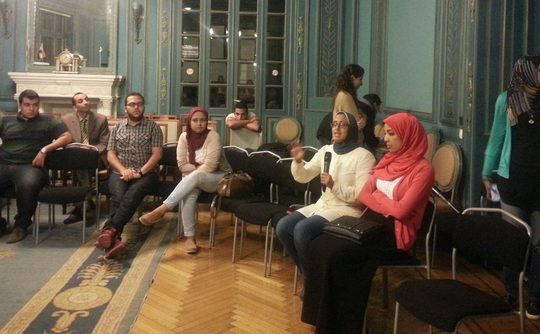 A group of people sat down in a large historical room with big windows, gold details on the walls. They are sat in a semi-circle, and one person is speaking and is holding a microphone.