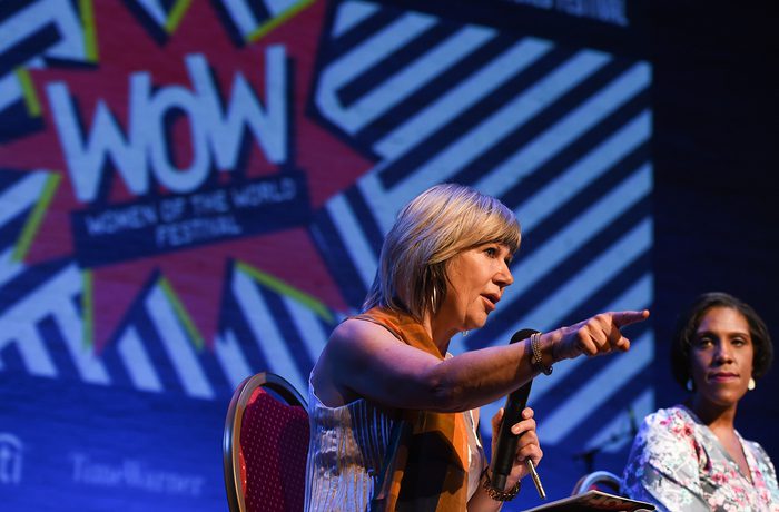 Two women on stage, in front of a screen which says "WOW women of the world festival". The women closest to the camera is speaking, holding a microphone, and pointing into the audience