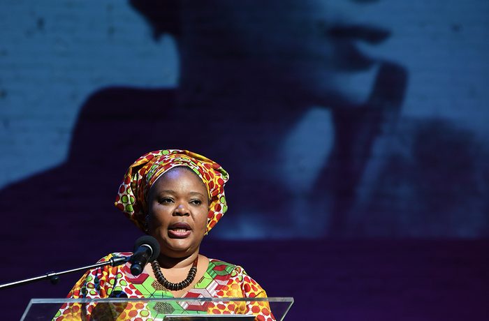 A woman stands on stage at a clear podium, speaking into a microphone. Behind her is a projected black-and-white photo of another woman looking off camera