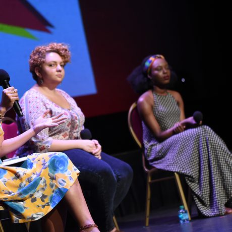 Four women sit on red chairs on stage, each holding a microphone. The woman nearest the camera is speaking and looking at the audience.
