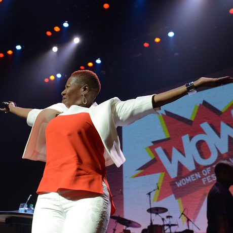 Woman performs on stage in white and red, arms outstretched in a victorious pose, with 'WOW Women of the World Festival' screen behind.