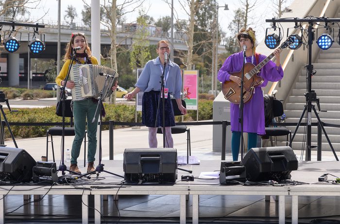Three women sing on an outdoor stage; one plays accordion, another plays guitar.
