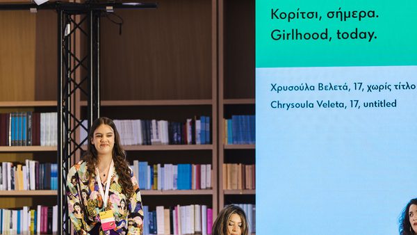 A young woman stands confidently on stage with a backdrop of bookshelves. Next to her, a large screen displays text about "Girlhood, today" in Greek and English.