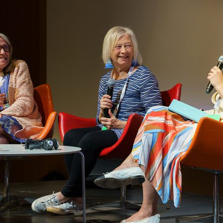 Three women sit on red chairs on stage with microphones; one speaks to the others. There is a table with water bottles in front