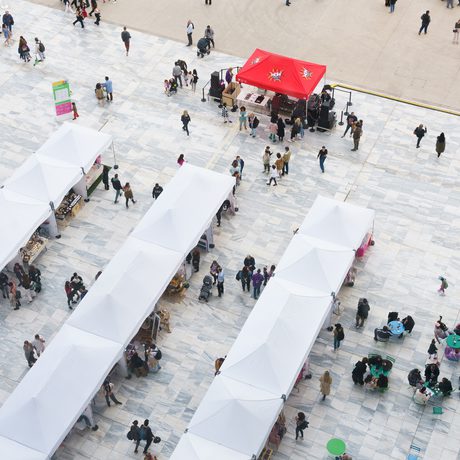 Aerial view of a busy market with white stalls; one bright red stall displays 'WOW Women of the World' logo.
