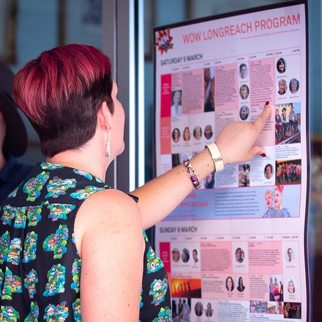 A person with short hair and a floral top points at a colourful poster on a glass door, discussing with two others.