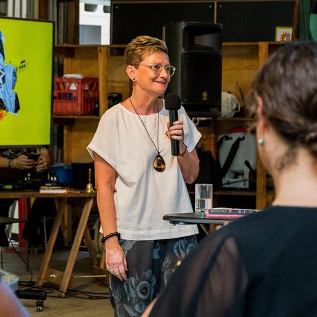 A woman with short hair speaks into a microphone, smiling, at a casual indoor event. Audience members listen, and a colourful screen displays WOW graphics behind her