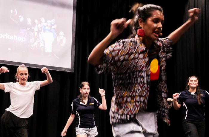 A group of four dancers energetically performing on stage, with a projected slide in the background.