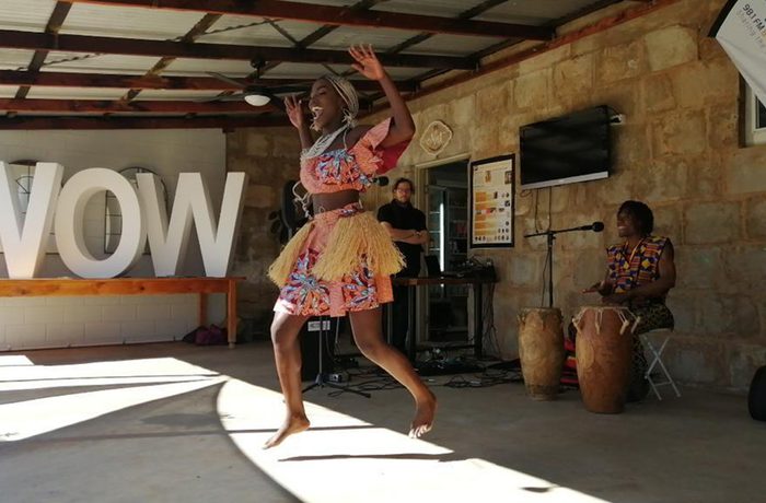 A dancer in colourful attire performs energetically on an outdoor stage with "WOW" in large letters. Musicians play drums behind her.