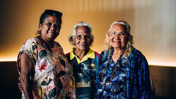Three older women stand together, smiling warmly. They wear colourful, patterned clothing against a softly lit background