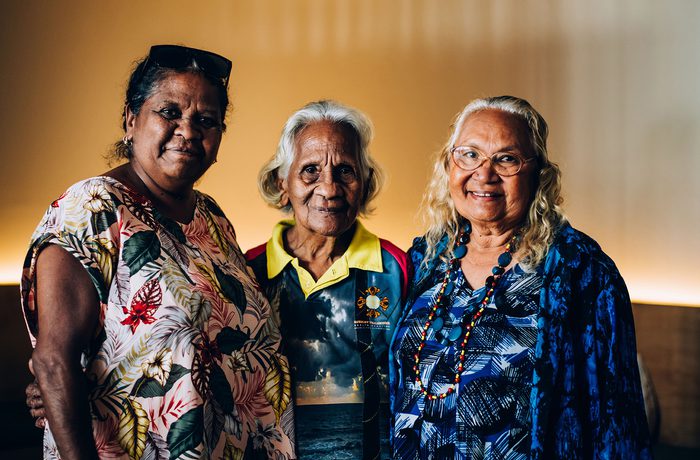Three older women stand together, smiling warmly. They wear colourful, patterned clothing against a softly lit background