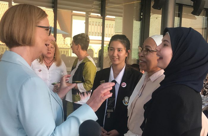 A woman in a light blue blazer engages with three young women, indoors. The mood is friendly and conversational.