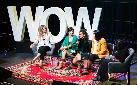 Five women sit on stage in conversation, framed by large "WOW" letters. They appear engaged and cheerful, seated on chairs over a patterned rug.